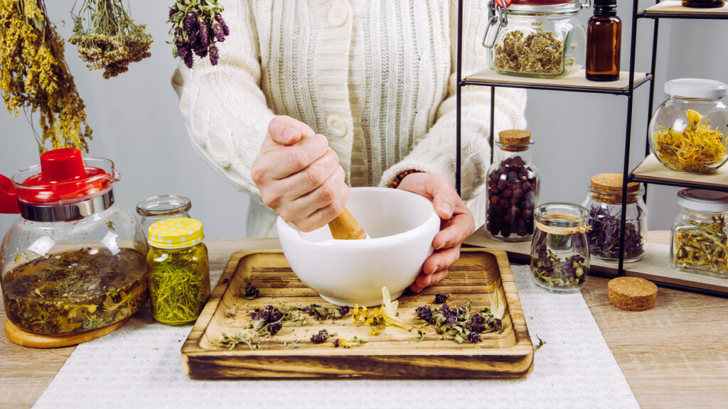 Close Up View Of Woman Herbalist Mixing Various Dried Herbs For Traditional Medicinal Tea With Mortar And Pestle. Dried Herbs In Glass Jars On Background.
