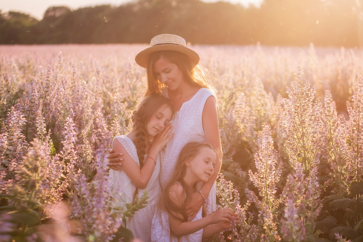 Young Mother Embraces Her Two Daughters Among A Purple Blooming Sage Field. Dressed In White With Straw Hats. Woman Lovingly Hugs Girls, Strokes, Kisses. Out Of Town Walks, Weekend Travel. Copy Space